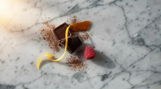 Composition elegante de carres de chocolat noir associes a des zestes d'agrumes, une framboise fraiche et des eclats de caramel sur un plan de travail en marbre, illustrant les harmonies gustatives de la patisserie francaise
